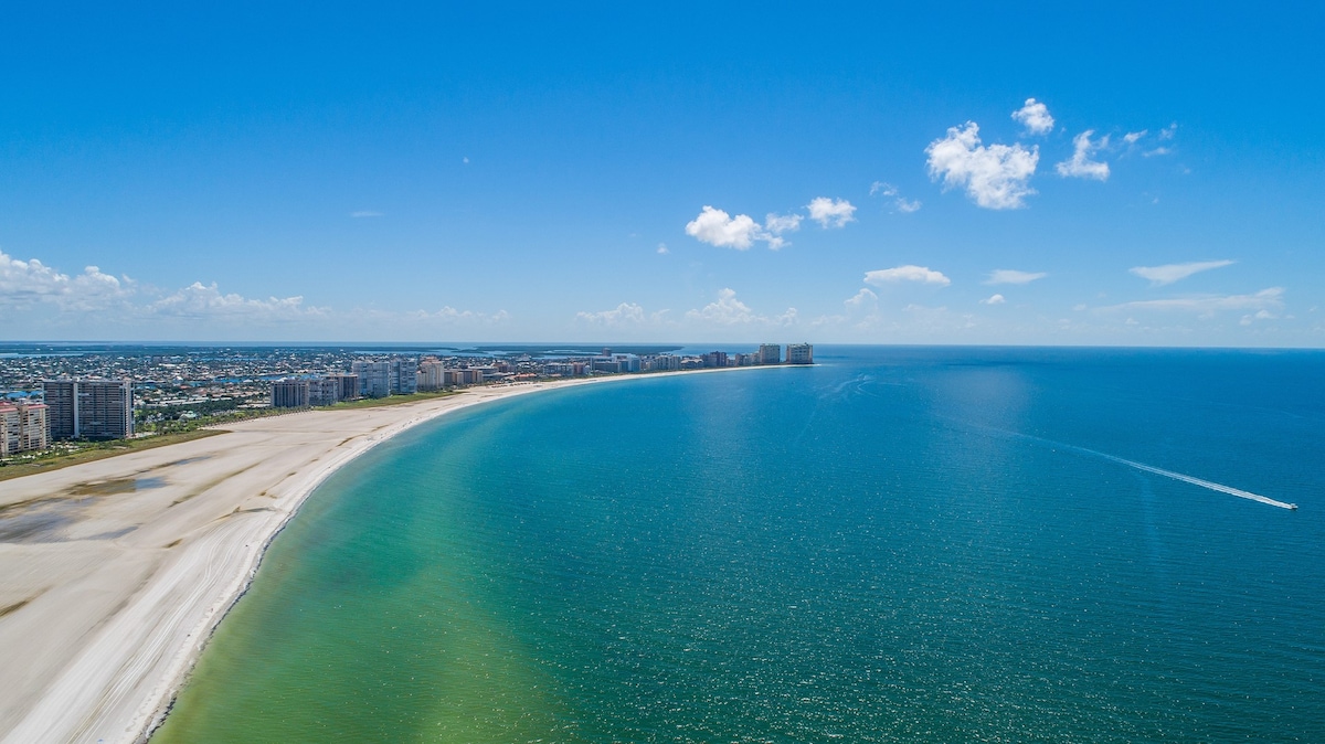An aerial view showcases the sweeping curve of Marco Island's white sand beach bordered by the shimmering aqua Gulf waters. The horizon stretches infinitely, with the coastline dotted by modern buildings under a bright blue sky, accentuated by wispy clouds.