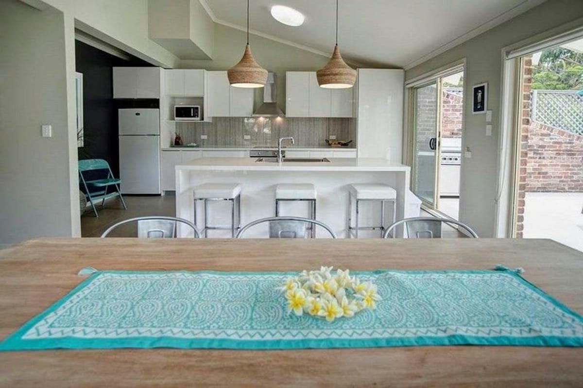 An inviting dining area features a wooden table adorned with a light blue decorative runner and a cluster of flowers. The adjoining kitchen is equipped with modern appliances, complemented by bar stools and pendant lighting, enhancing the open plan design.