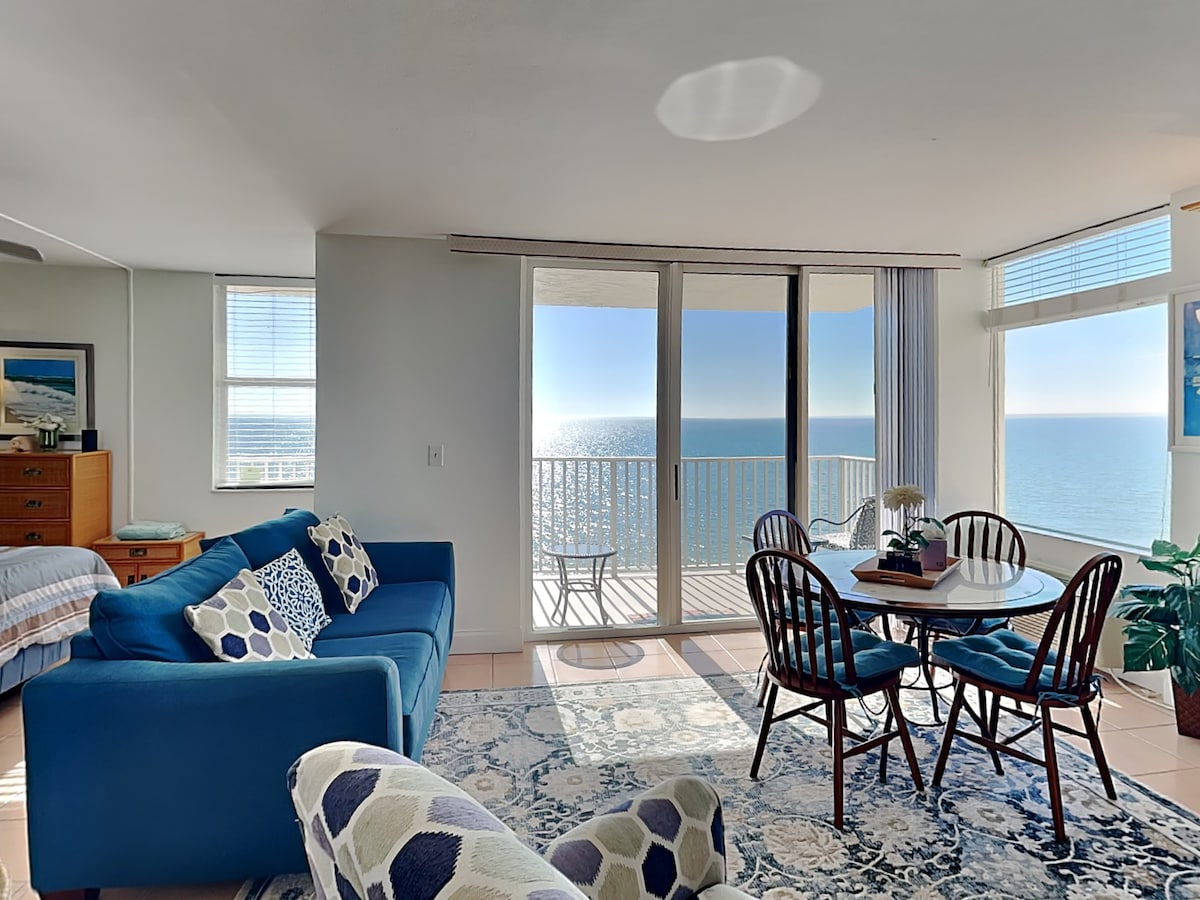 Natural light fills an open living and dining area, featuring a blue sofa and a round dining table set for four. The sliding glass doors reveal expansive views of the Gulf, complemented by a small balcony with seating. Soft area rugs enhance the inviting space.