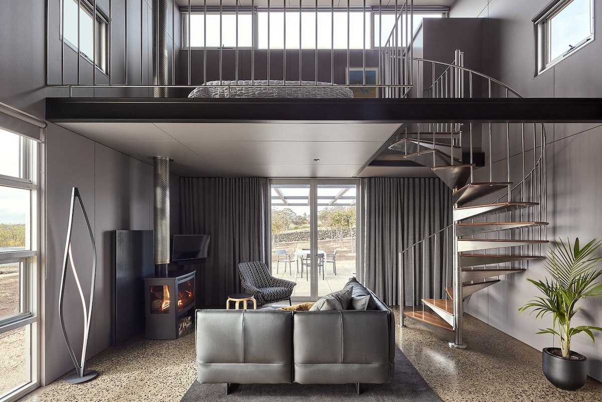 A split-level interior features a cozy seating area with a sleek grey couch facing a modern wood stove. A spiral staircase leads to a mezzanine sleeping space. Large windows frame views of the surrounding landscape, while dark curtains add a touch of elegance.