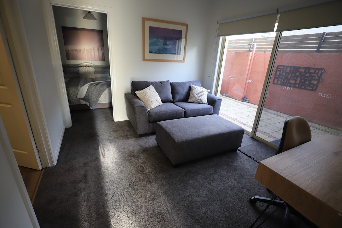 A cozy living area is furnished with a grey sofa and ottoman, accompanied by a wooden desk. Natural light enters through sliding glass doors that offer a view of the private courtyard. The space features soft carpeting, creating a comfortable atmosphere.
