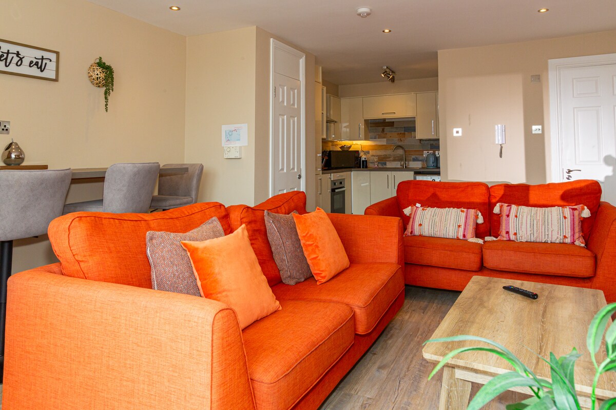 A spacious living area features bright burnt orange sofas adorned with decorative pillows. A wooden coffee table sits in front, while a modern kitchen area is visible in the background. Dining options are available at a bar with high stools along the wall.
