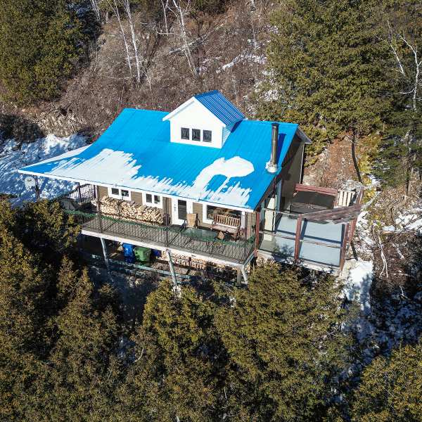 An aerial view of the cottage showcases a blue metal roof and a wraparound porch surrounded by greenery. The structure is partially obscured by trees, providing a sense of privacy. Snow can be seen on the roof, indicating the seasonal setting of the property.