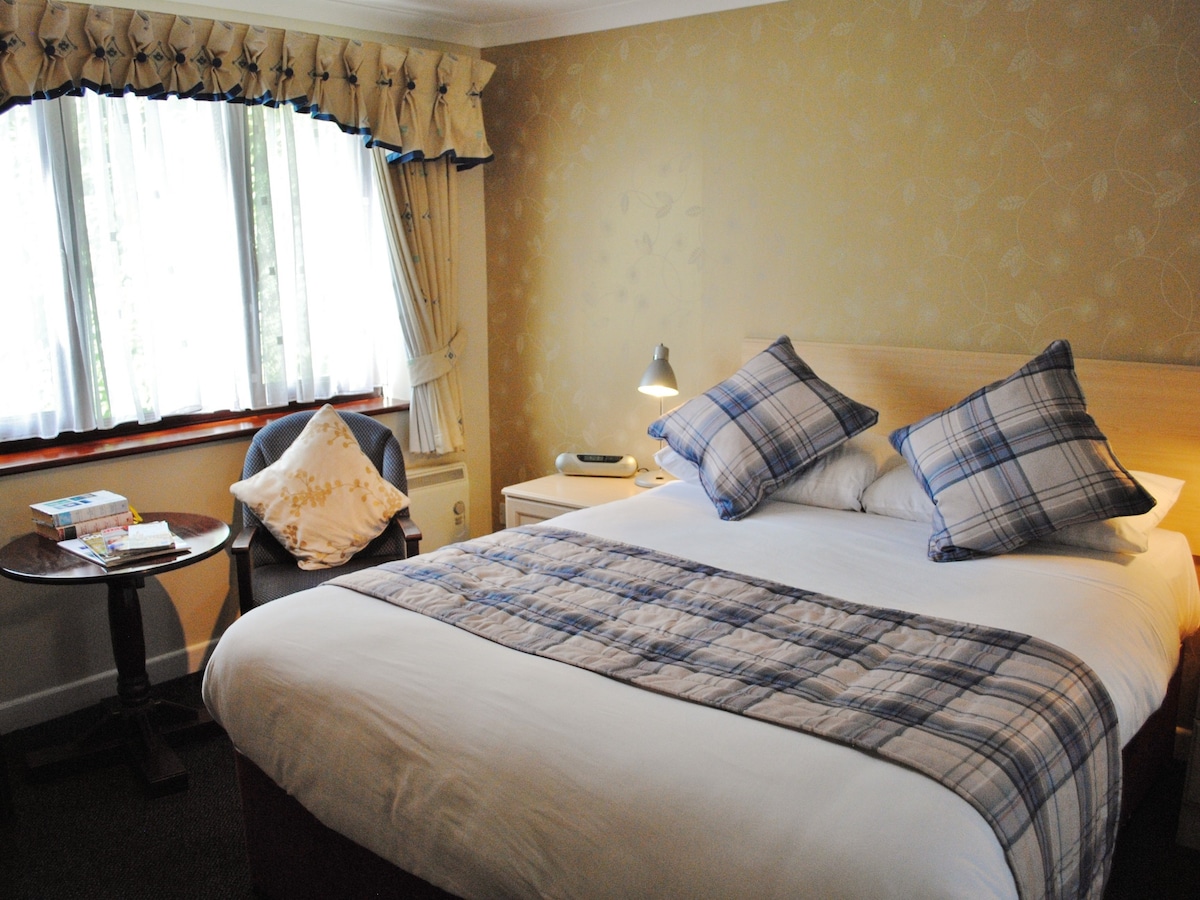 A well-lit bedroom features a comfortable double bed with a soft quilt and plaid accent pillows. A small round table and a chair are positioned near a large window, which is dressed with light drapes. A stack of books adds a cozy touch to the room.