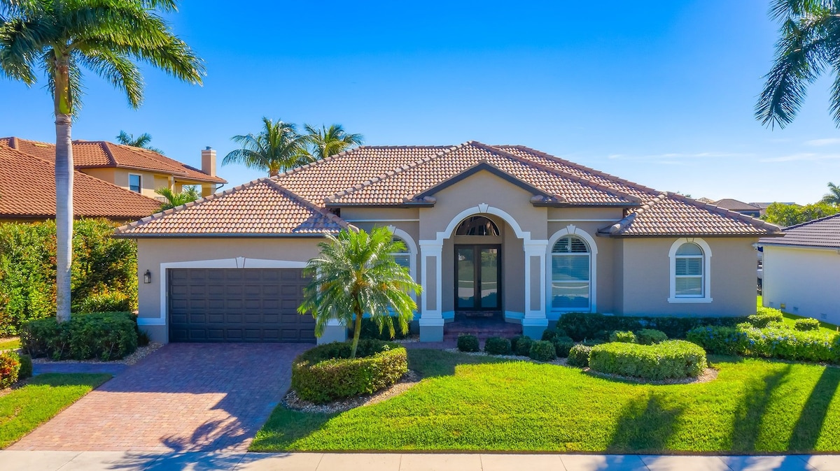 A spacious waterfront home is depicted with a decorative tile roof and an elegant entrance. Lush landscaping surrounds the property, featuring palm trees and manicured shrubs. A paved driveway leads to a two-car garage, under a clear blue sky.
