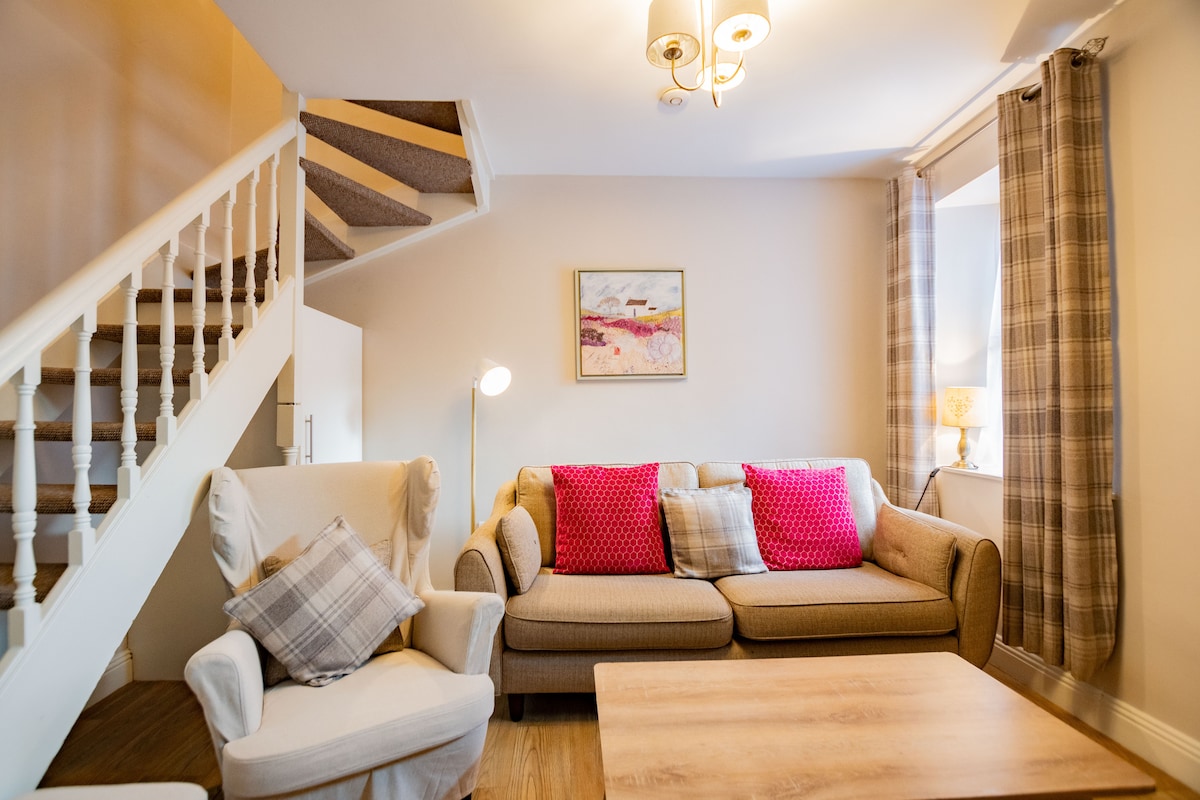 A comfortable living area features a sofa adorned with throw pillows in vibrant pink and neutral tones. A wooden coffee table is positioned centrally, and warm lighting is provided by a stylish lamp. Stairs lead to the upper level, framed by soft curtains at the window.
