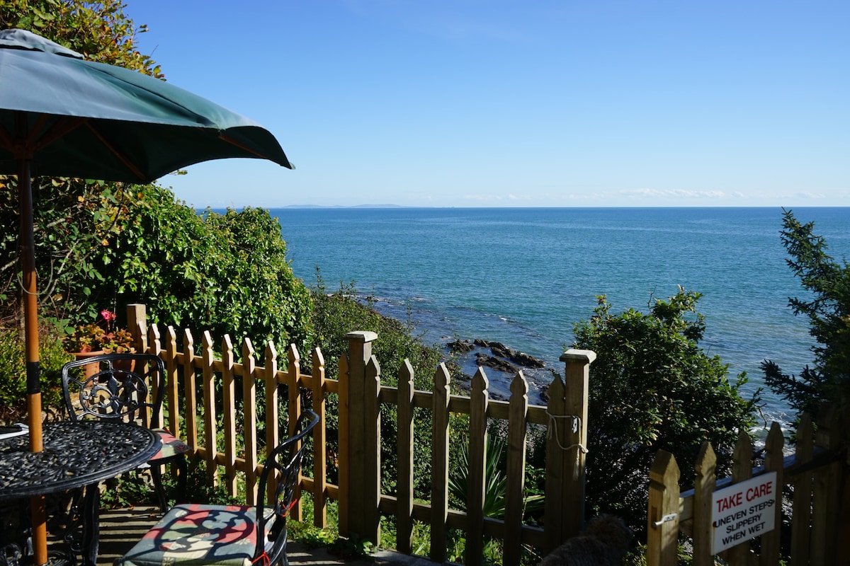 A private outdoor seating area features a round table and chairs, framed by a wooden fence. The clear view of the sea extends to the horizon, while lush greenery and coastal rocks are visible in the foreground under a bright blue sky.