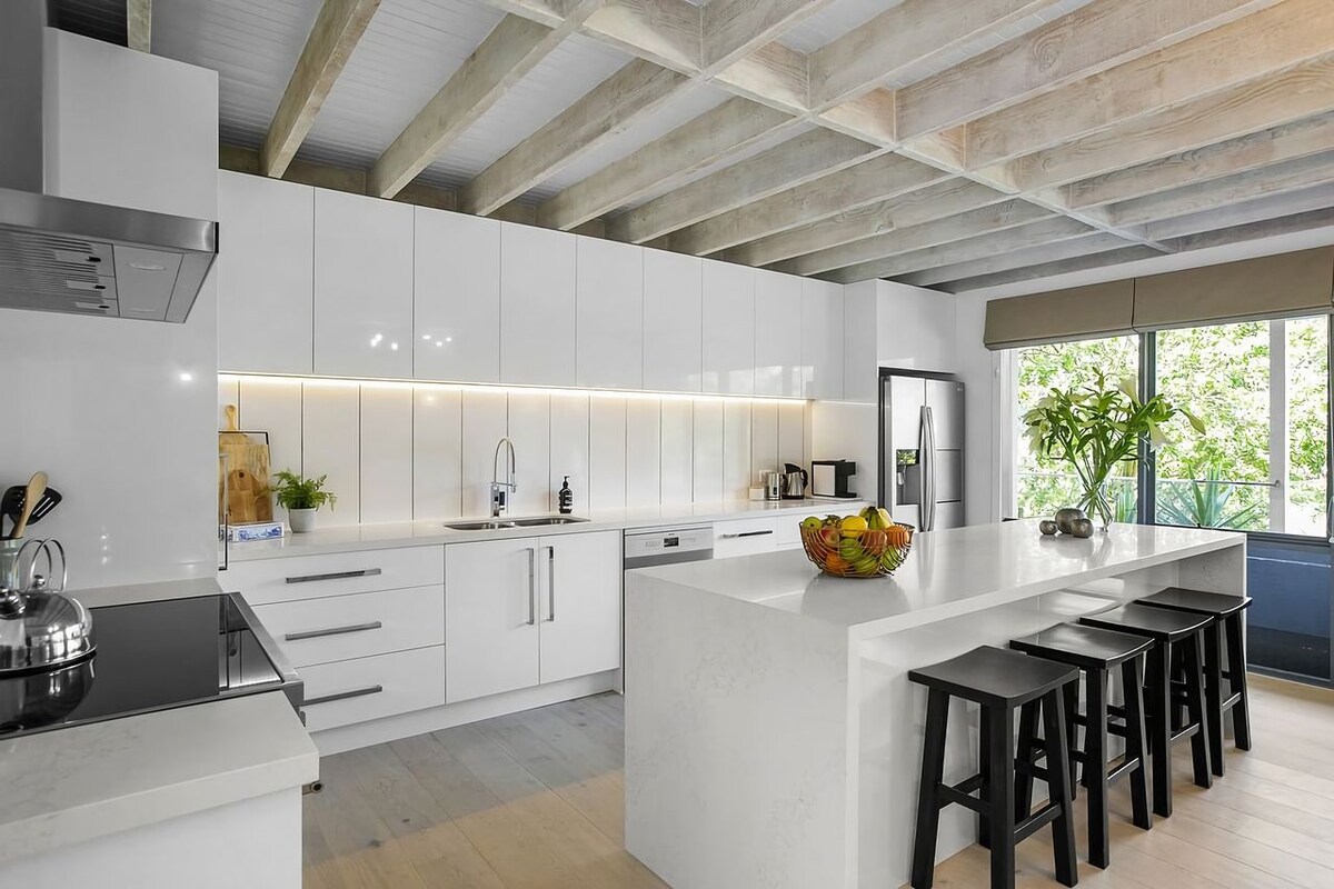 A modern kitchen is showcased with sleek white cabinetry and polished stone countertops. A large island with seating is complemented by a bowl of fresh fruit. Natural light filters through wide windows, enhancing the airy feel of the space.