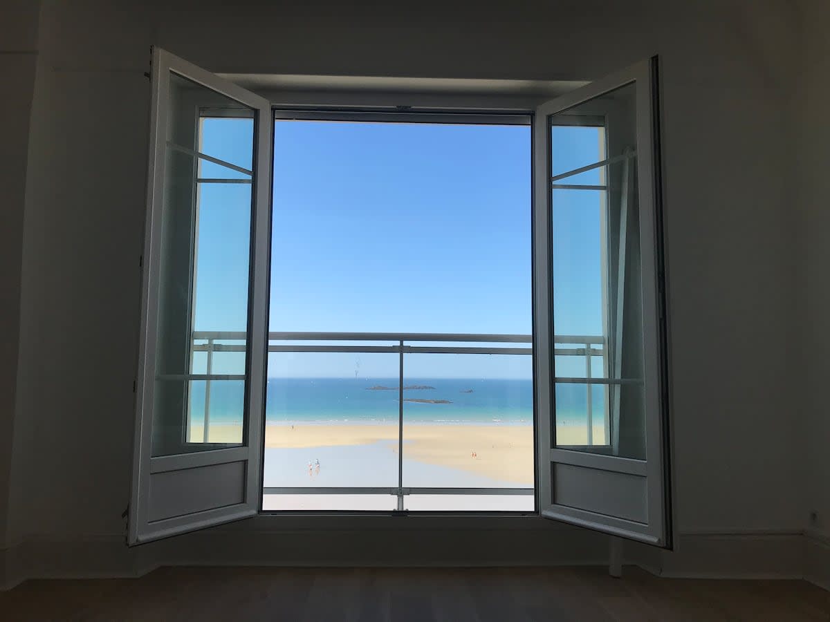 Large open windows frame a breathtaking view of the sandy beach and clear blue sea. The scene captures a bright sky with minimal cloud coverage, inviting natural light into the space.