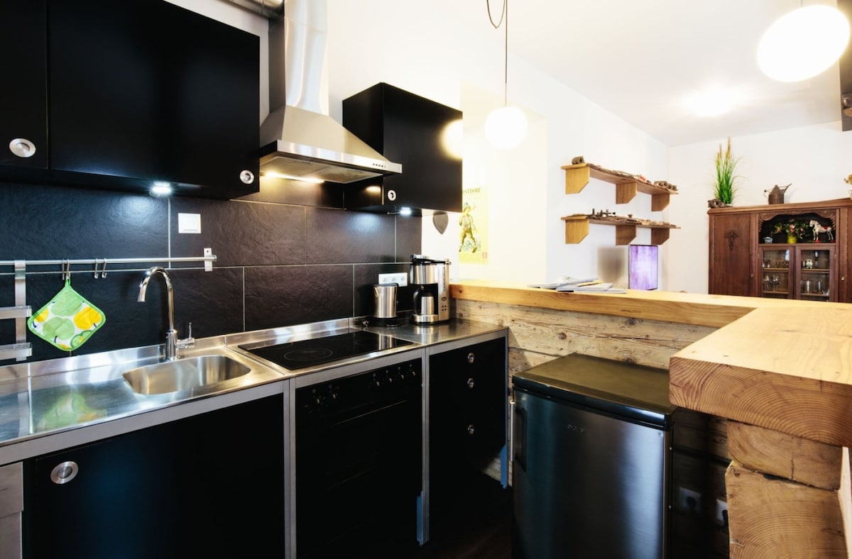 A well-equipped kitchen is designed with modern black cabinetry and a stainless steel refrigerator. An open breakfast bar with a wooden countertop provides additional seating. Stainless steel appliances, including an oven and a dishwasher, are integrated within the space, accompanied by under-cabinet lighting.