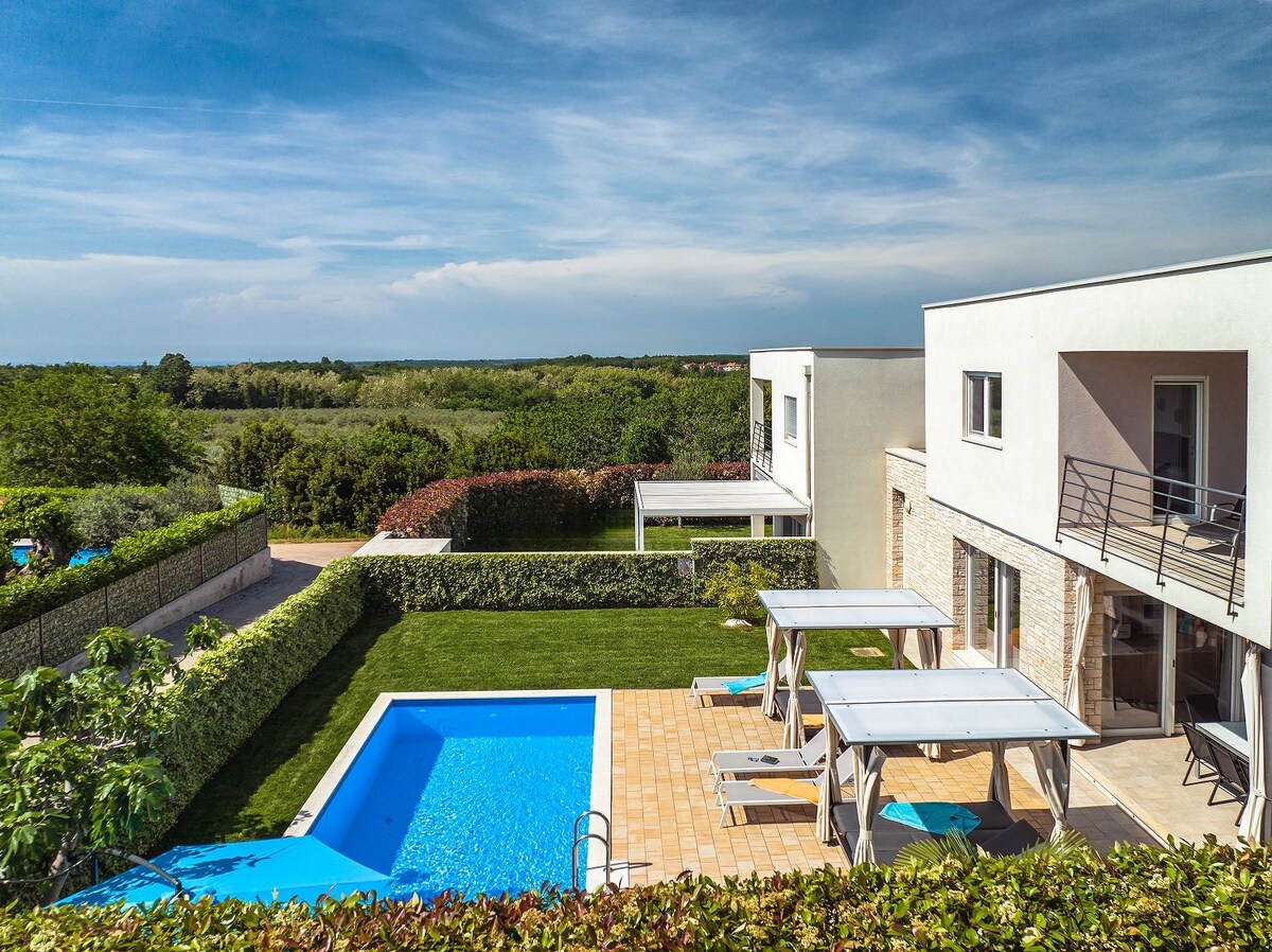 A spacious outdoor area is visible, featuring a private heated pool surrounded by a landscaped lawn. Sun loungers are positioned near the pool, and a covered seating area is included. The villa has modern architecture with large windows, and lush greenery can be seen in the background.