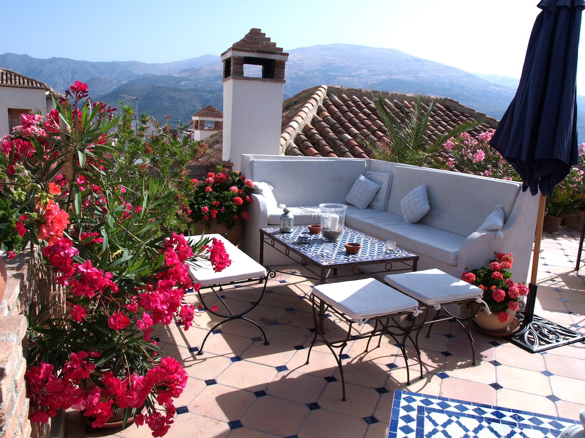A roof terrace is adorned with vibrant flower pots, featuring comfortable seating arrangements around a glass-topped table. The space is designed for relaxation, with scenic mountain views visible in the background. Sun loungers are placed nearby on the tiled surface.