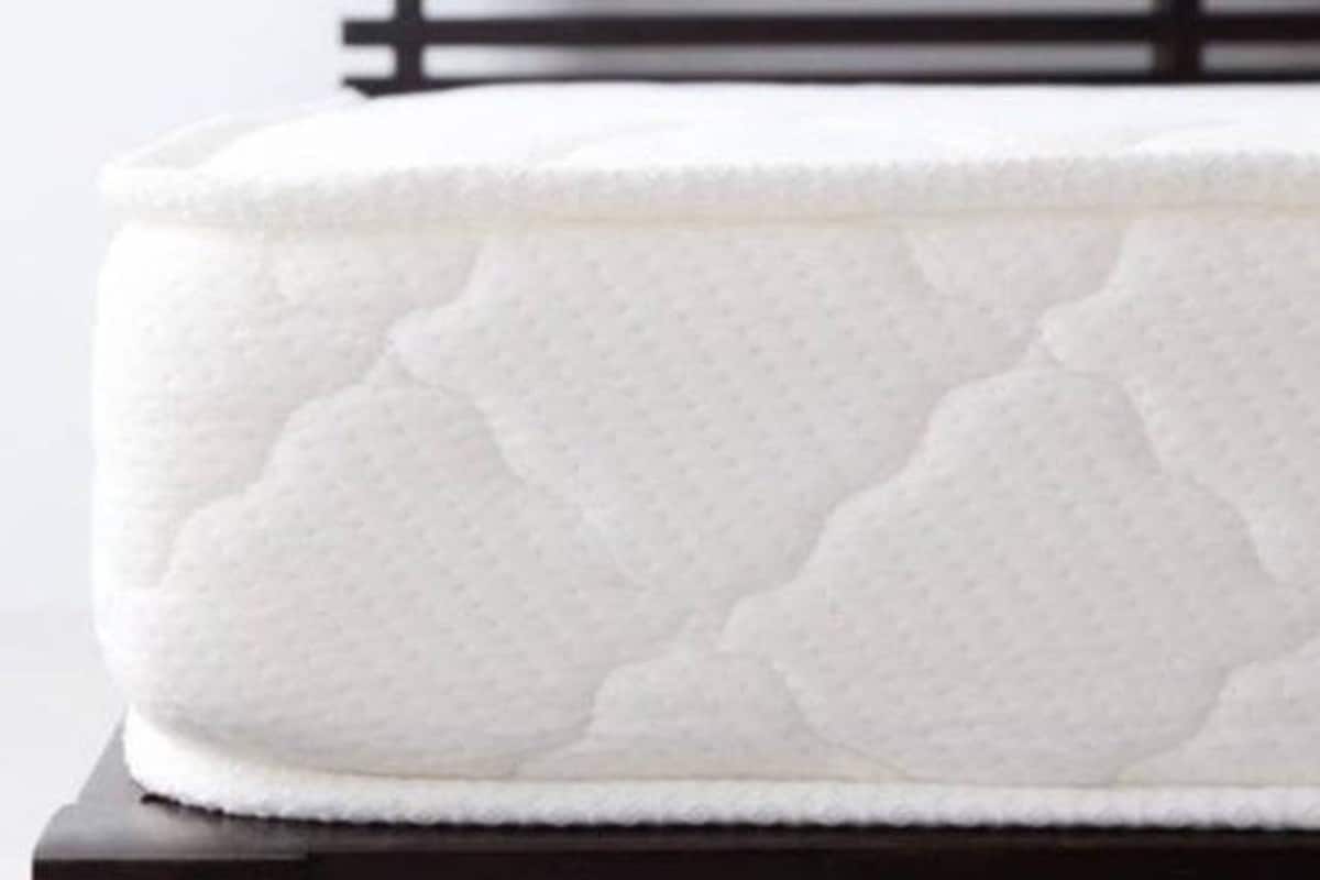 A close-up image of a queen-size mattress is displayed, showcasing its soft, quilted surface. The edges are neatly finished, and it is positioned on a wooden bed frame. The color is primarily white, indicating cleanliness and comfort.