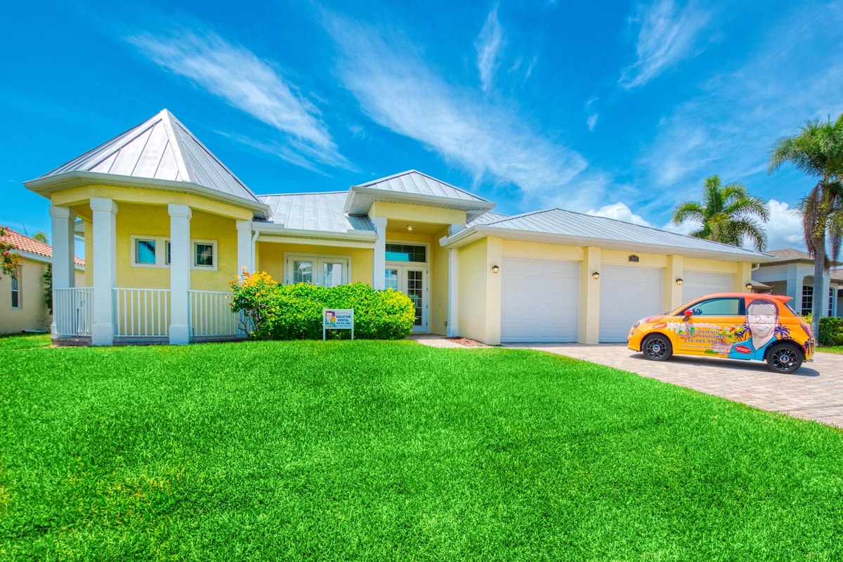 The exterior of a vibrant yellow home is showcased against a clear blue sky. A neatly manicured lawn surrounds the entrance, which features a covered porch. In the driveway, a small, colorful car is parked, enhancing the welcoming appeal of the property.
