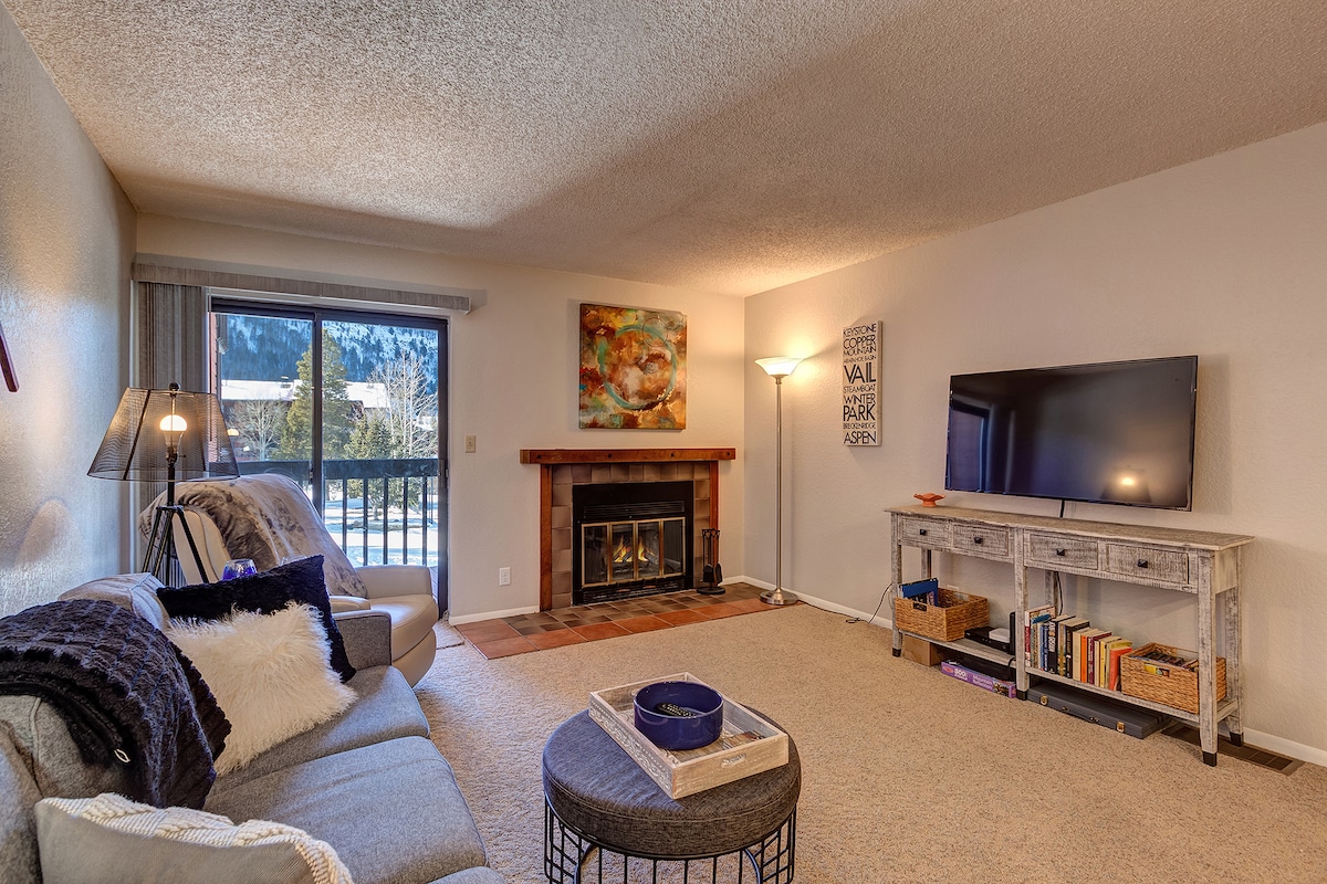 A comfortable living area features a plush sofa and an armchair, positioned around a coffee table. A flat-screen TV is mounted on the wall, while a decorative fireplace adds warmth. Natural light enters through sliding glass doors leading to an outdoor space with mountain views.