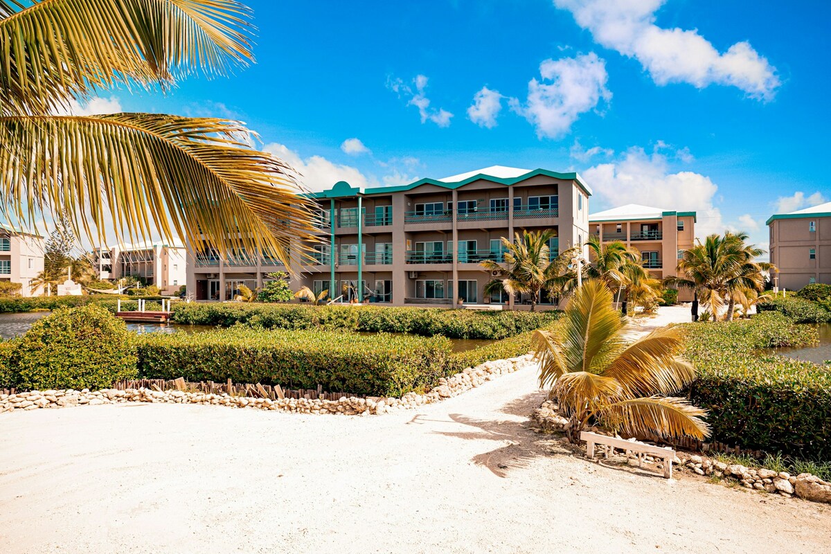 A tropical landscape in front of the suite showcases lush greenery and palm trees. The three-story building features balconies, allowing for outdoor relaxation. A sandy pathway leads toward the water, creating an inviting atmosphere. Clear blue skies enhance the overall brightness of the scene.