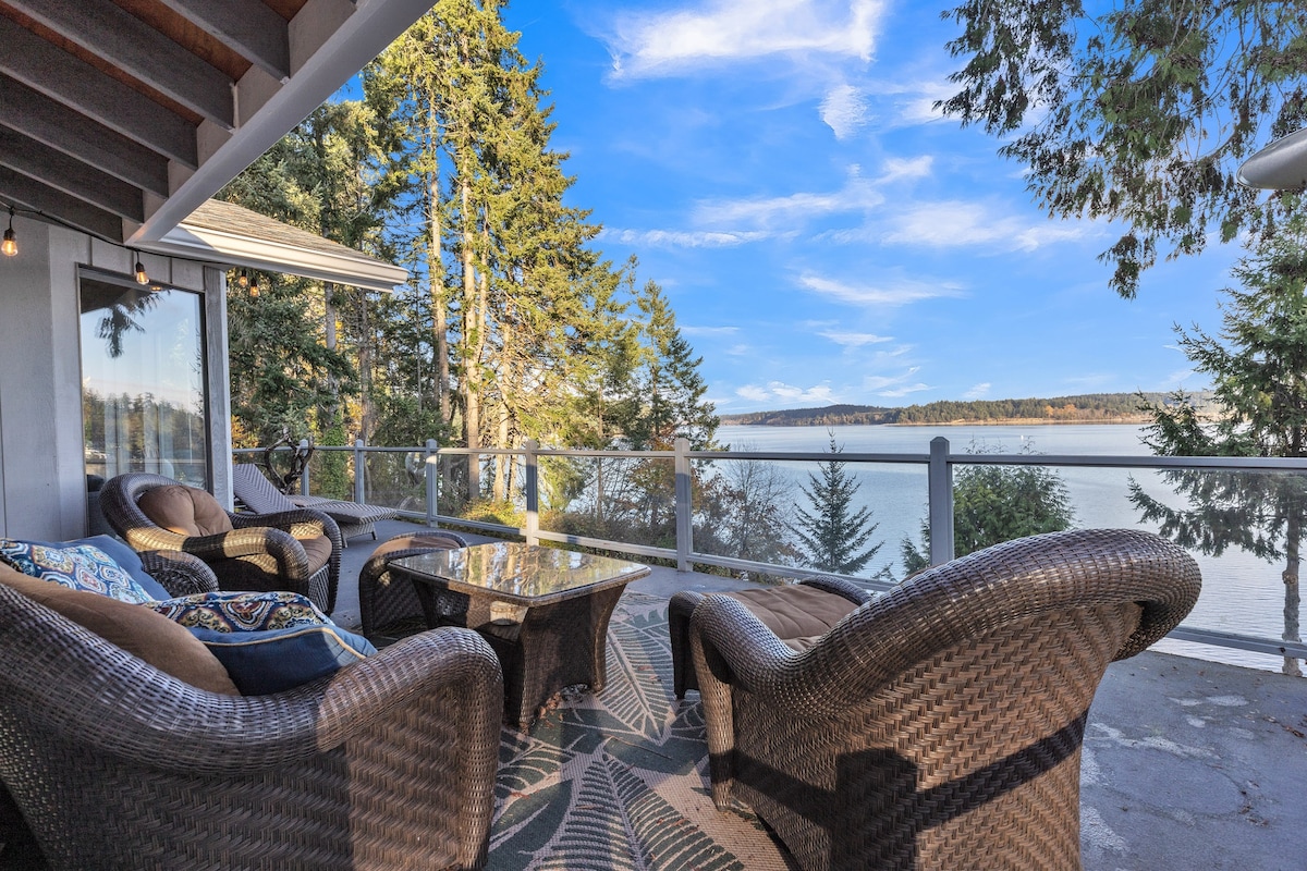 A spacious deck is furnished with comfortable seating arrangements, including wicker chairs and a glass-top table. The view encompasses a serene waterfront, framed by lush trees, with distant land visible across the bay under a clear blue sky.
