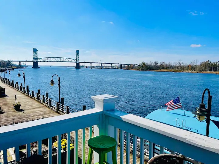 The image captures a scenic view from a balcony overlooking the Cape Fear River. The calm water reflects the blue sky, while a bridge spans the river in the distance. A green stool and lamp are positioned on the railing, enhancing the outdoor space.