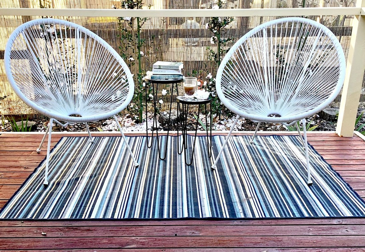 A cozy outdoor seating area features two white woven chairs positioned on a striped rug. A small table holds a book and a drink, surrounded by greenery and a light wooden railing, creating an inviting space for relaxation.