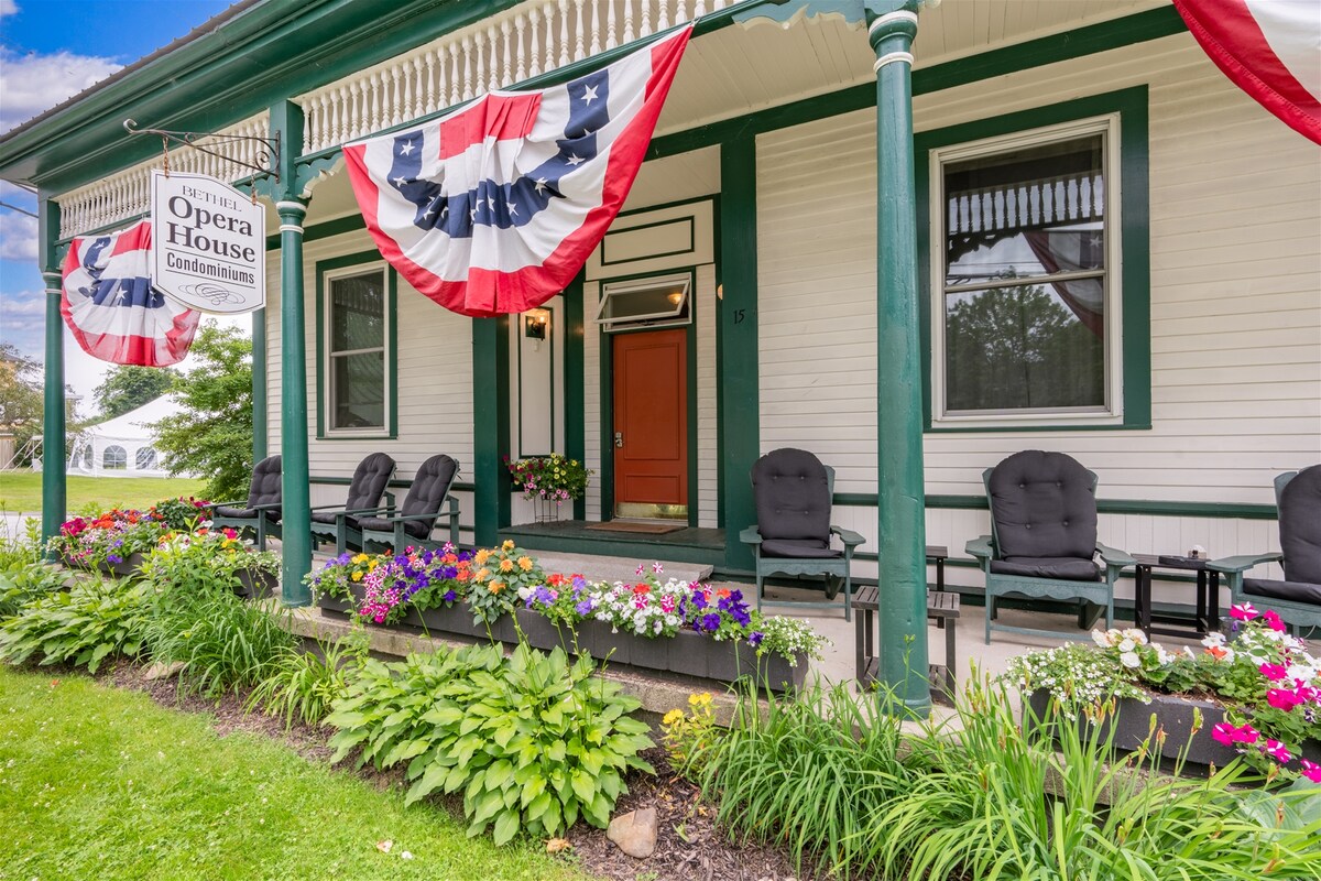 A charming front porch is adorned with black rocking chairs and vibrant flower beds, creating a welcoming entryway. Decorative red, white, and blue bunting hangs from the porch, adding a festive touch. The inviting entrance features a bright orange door that opens to the inside.
