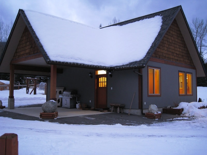 The cabin features a sloped roof with a combination of shingle and painted siding, set against a snowy landscape. A covered entryway is visible, with warm light spilling from windows. A deluxe gas grill is situated to the side, complemented by natural stone elements in the foreground.