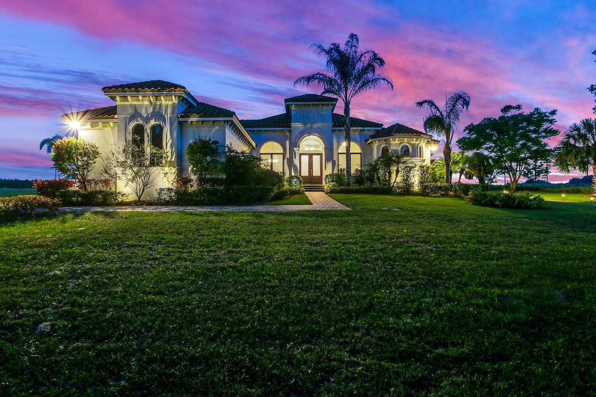 A luxurious mansion is displayed against a vibrant sunset sky, highlighting its elegant architectural features. Lush greenery and palm trees frame the property, while soft lighting accentuates the entryway and pathways, creating an inviting ambiance at dusk.