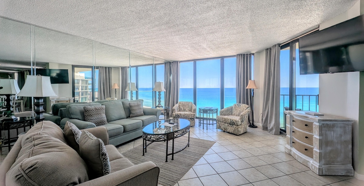 A spacious living area is defined by large windows providing panoramic views of the ocean. The area features comfortable seating arrangements with a sofa and armchairs, a stylish glass coffee table, and a media console with a television. Natural light enhances the serene ambiance.