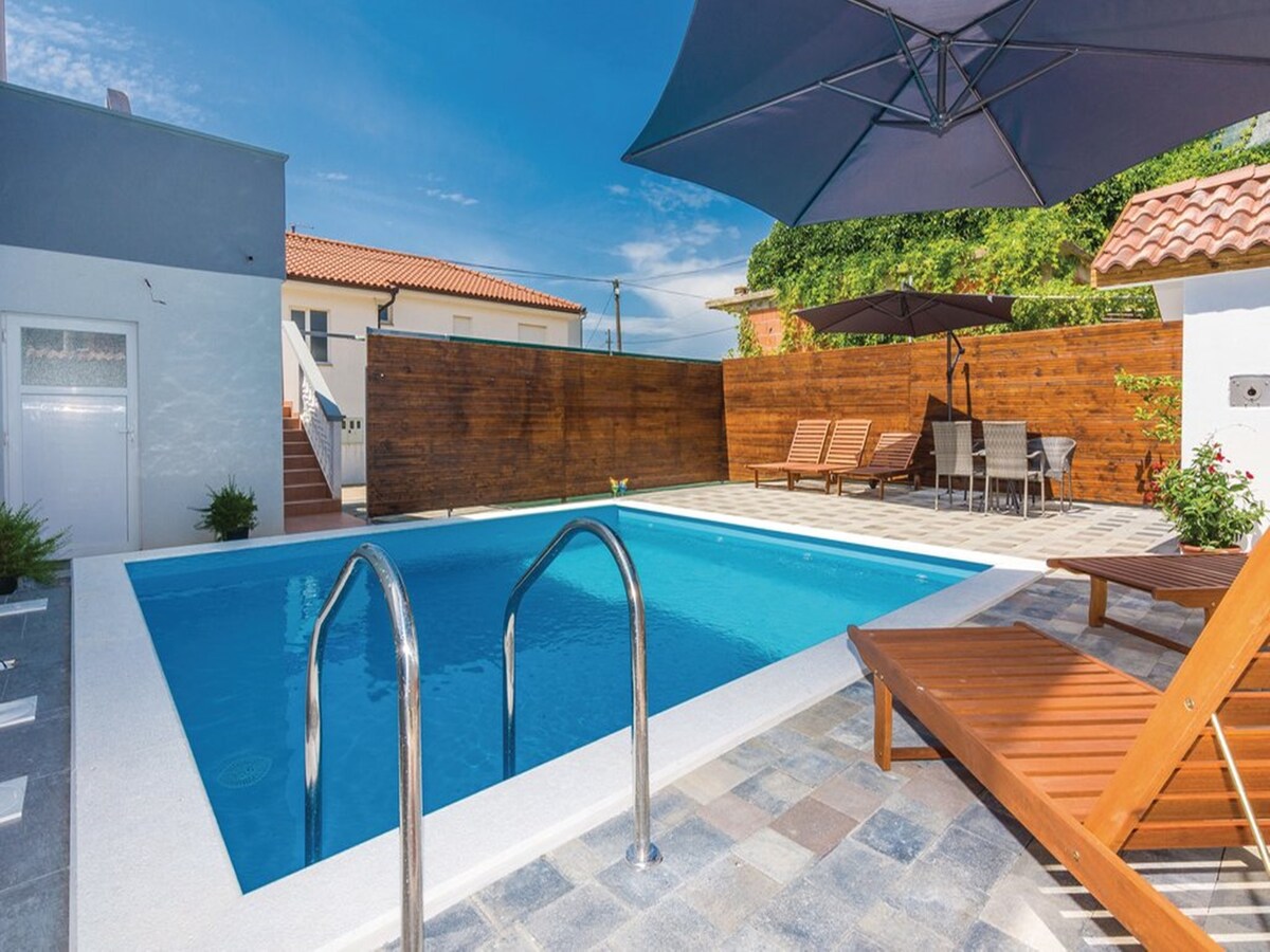 A private outdoor area features a clear swimming pool surrounded by a tiled deck. Comfortable sun loungers are positioned nearby, and a large umbrella provides shade. Wooden fencing surrounds the space, offering privacy, with a dining area visible in the background.