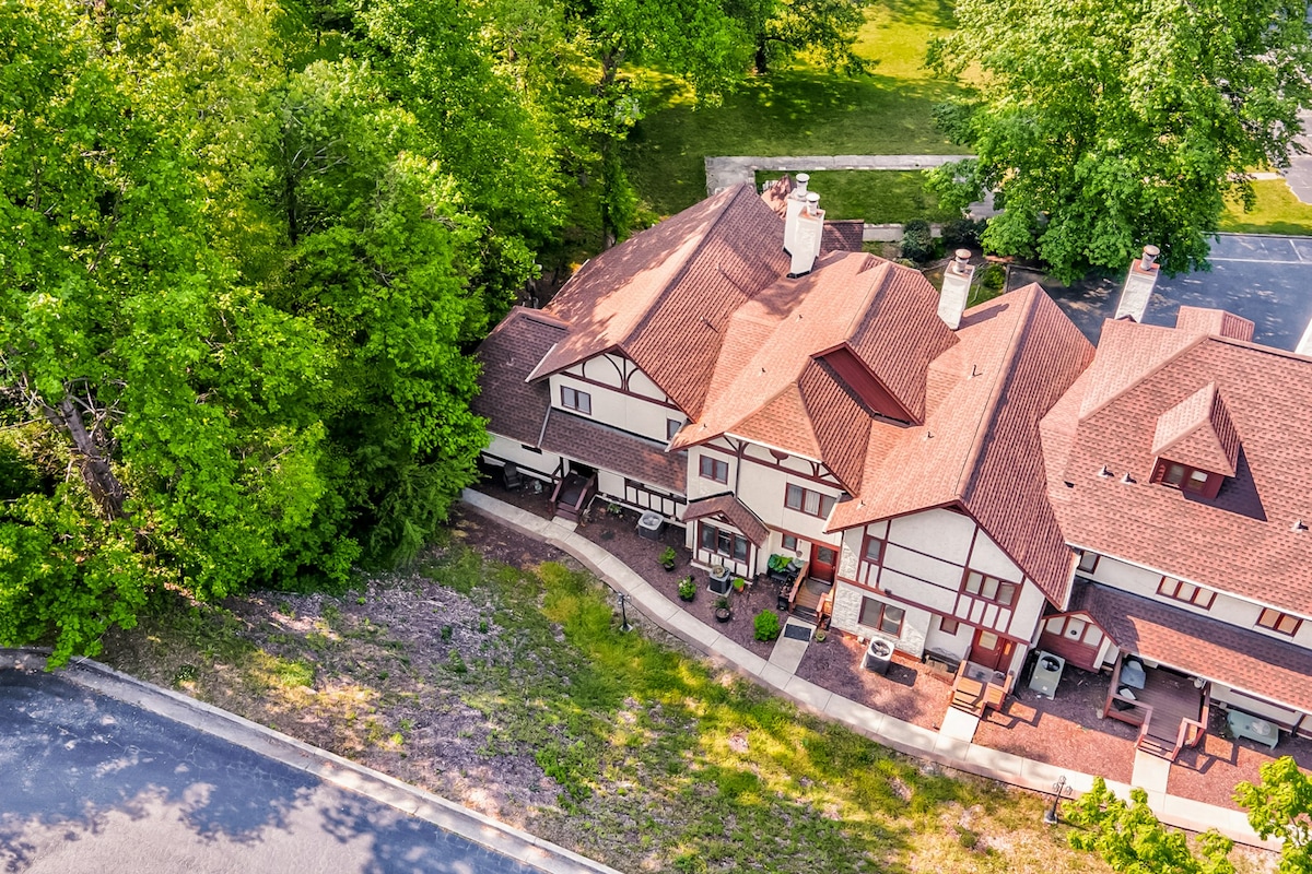 An aerial view of a multi-level condo surrounded by lush greenery and trees. The building features a distinctive architectural style with a terracotta shingle roof and white siding. The outdoor area is adorned with planters and a paved walkway leading to the entrance.