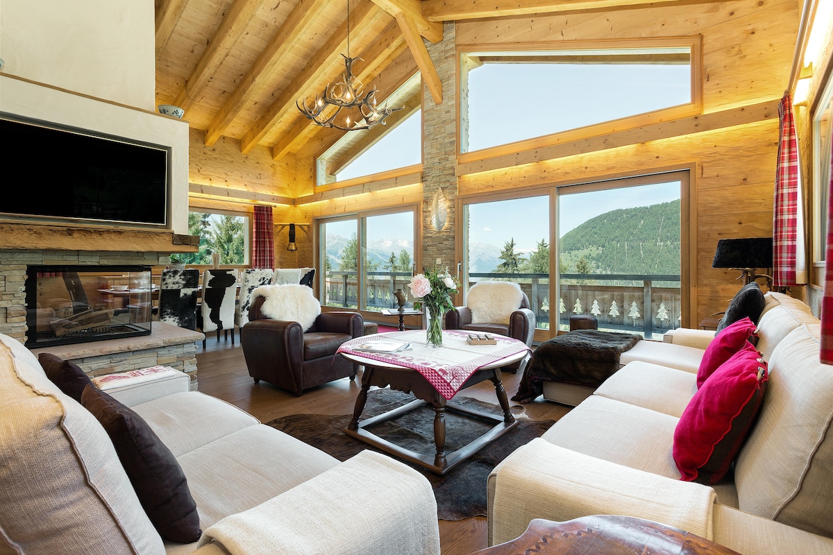 A spacious living area features a high, wooden-beamed ceiling and large windows that frame scenic views of the surrounding mountains. Comfortable sofas in soft colors are arranged around a central coffee table, complemented by decorative pillows in rich hues. A modern fireplace adds warmth to the atmosphere.