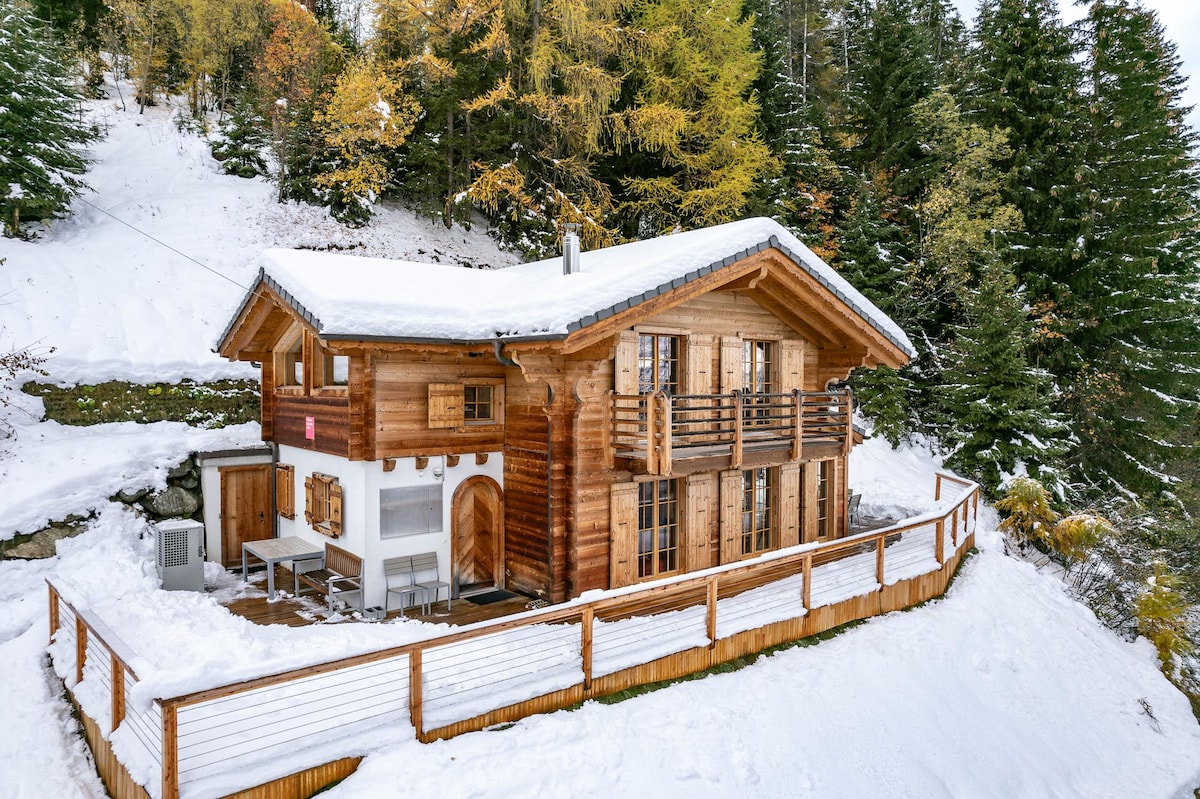 A charming wooden chalet is surrounded by snow-covered terrain, featuring multiple balconies with wooden railings. The stone foundation complements the warm wood finish, while the roof is blanketed in fresh snow. Tall evergreen trees border the chalet, adding to the serene winter landscape.