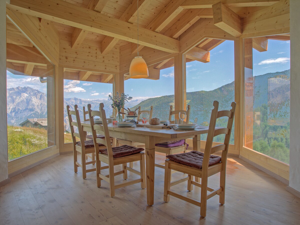 A spacious dining area features a wooden table surrounded by six chairs, with views of the mountains visible through large, floor-to-ceiling windows. Natural light fills the space, complemented by a hanging light fixture above the table.