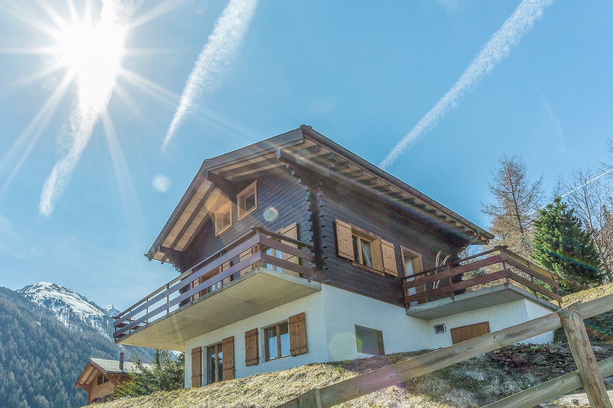 The chalet, constructed from larch wood, is presented from an outdoor perspective under a clear blue sky. Sunlight reflects off the structure, showcasing the balcony and large windows. Surrounding greenery and distant snow-capped mountains enhance the peaceful mountain setting.