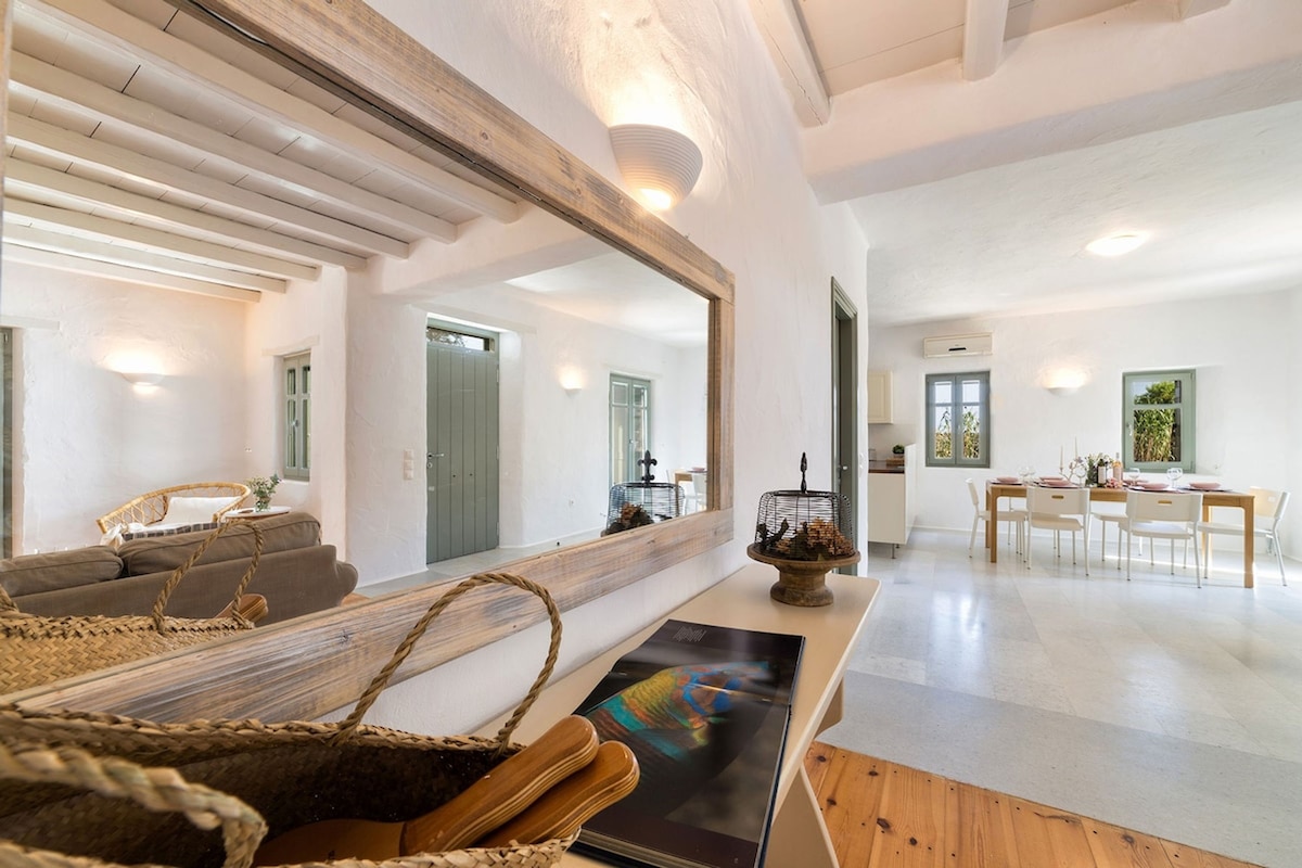 A light-filled space is shown with whitewashed walls and wooden beams on the ceiling. A mirrored wall reflects the open living area, which features a comfortable seating arrangement and a dining space in the background. Natural materials and soft lighting create a welcoming atmosphere.