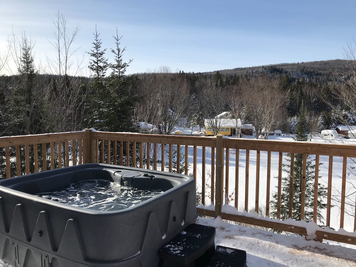 A private spa is situated on a wooden deck, surrounded by a wooden railing that offers views of the snow-covered landscape. Evergreen trees are visible in the background, contrasting with the clear blue sky overhead.