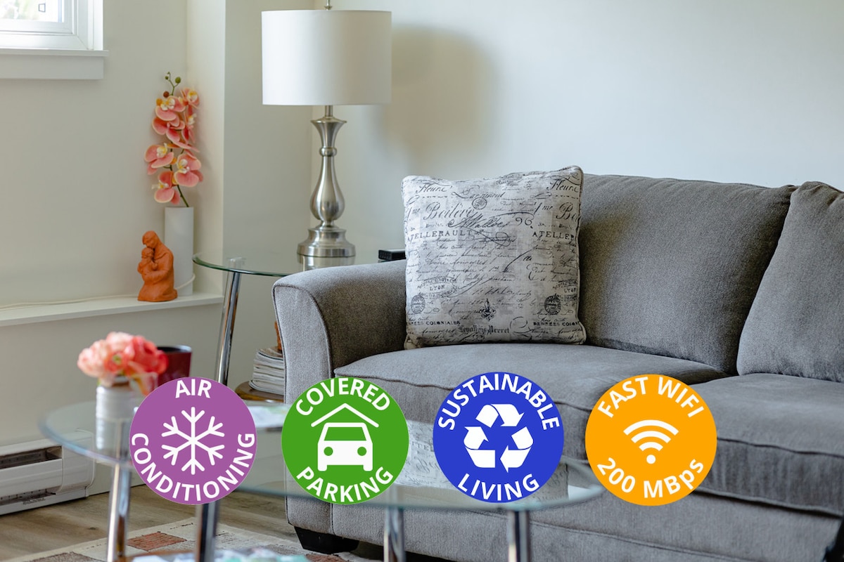 A comfortable gray sofa is positioned in the living area next to a glass-topped side table. Decorative elements include a small flower arrangement and a textured cushion. Informational icons signify air conditioning, covered parking, sustainable living, and high-speed Wi-Fi.