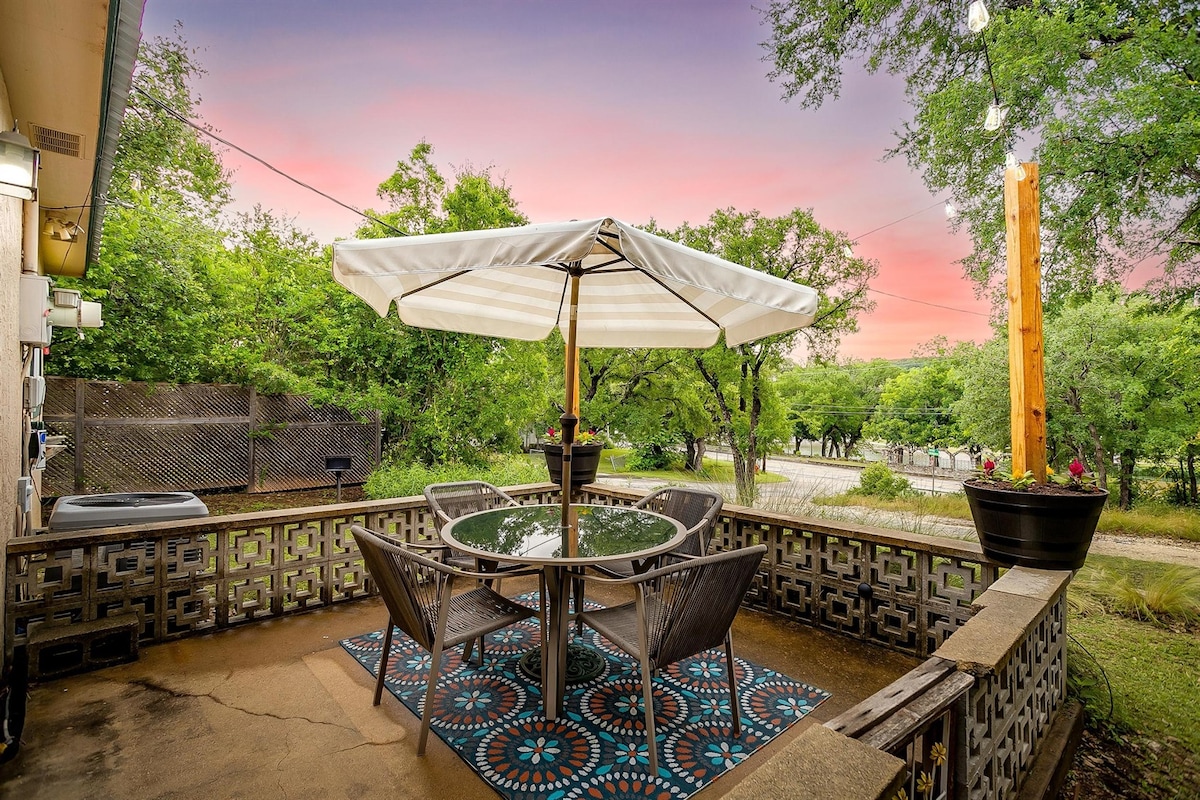 An outdoor patio features a round glass table surrounded by four chairs, beneath a large umbrella for shade. The area is adorned with a colorful patterned rug, while greenery surrounds the space, providing a serene view of the landscaping and nearby trees.