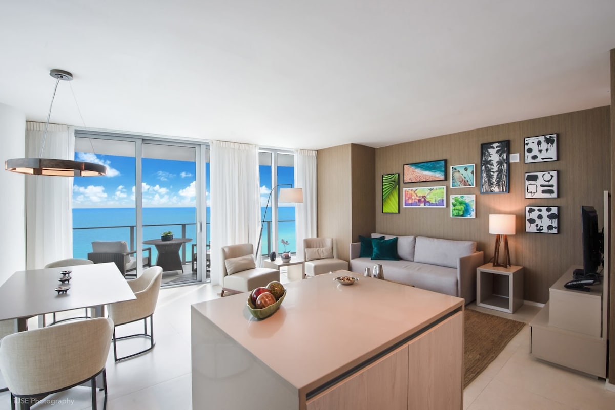 A modern living space features a comfortable sofa and side tables, alongside a dining area with a round table and upholstered chairs. Large glass doors open to a panoramic ocean view, while colorful wall art adds a touch of vibrancy to the neutral decor.