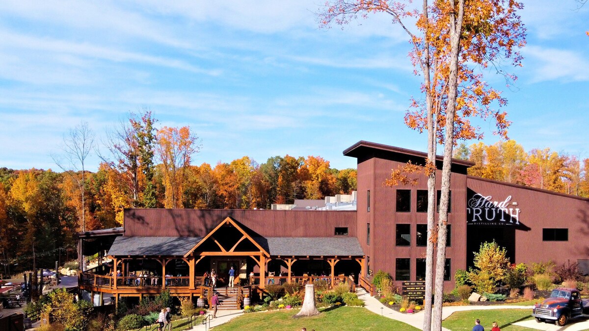 A modern building with wooden accents is set against a backdrop of autumn-colored trees. The structure includes a spacious deck with guests enjoying the outdoor seating, and a large sign that reads 'Find Truth' is prominently displayed on the facade.