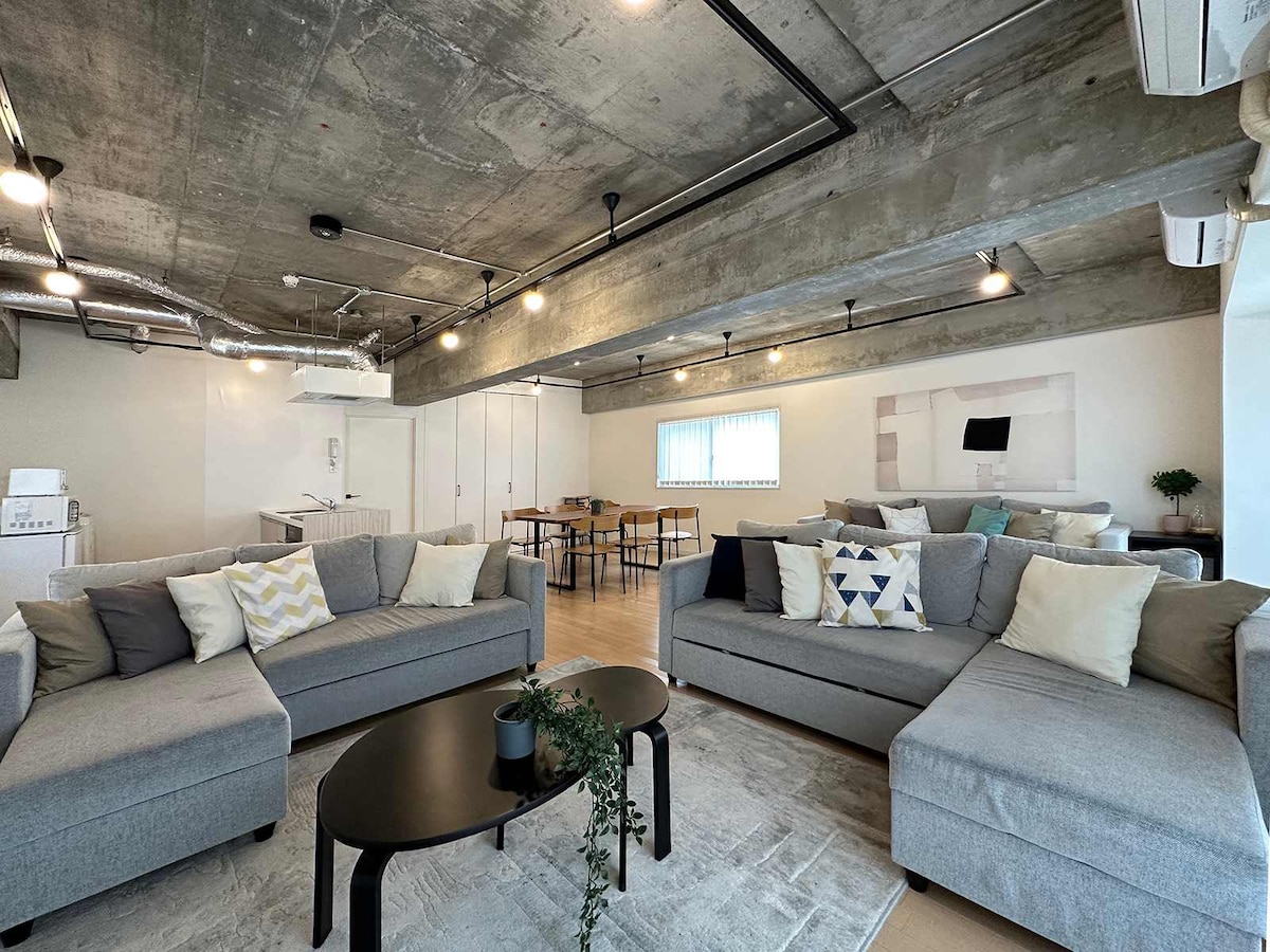 A spacious living area features comfortable gray sofas adorned with decorative pillows. A low black table sits at the center, complemented by a light-colored area rug. Large windows allow natural light to fill the space, enhancing the modern industrial design.