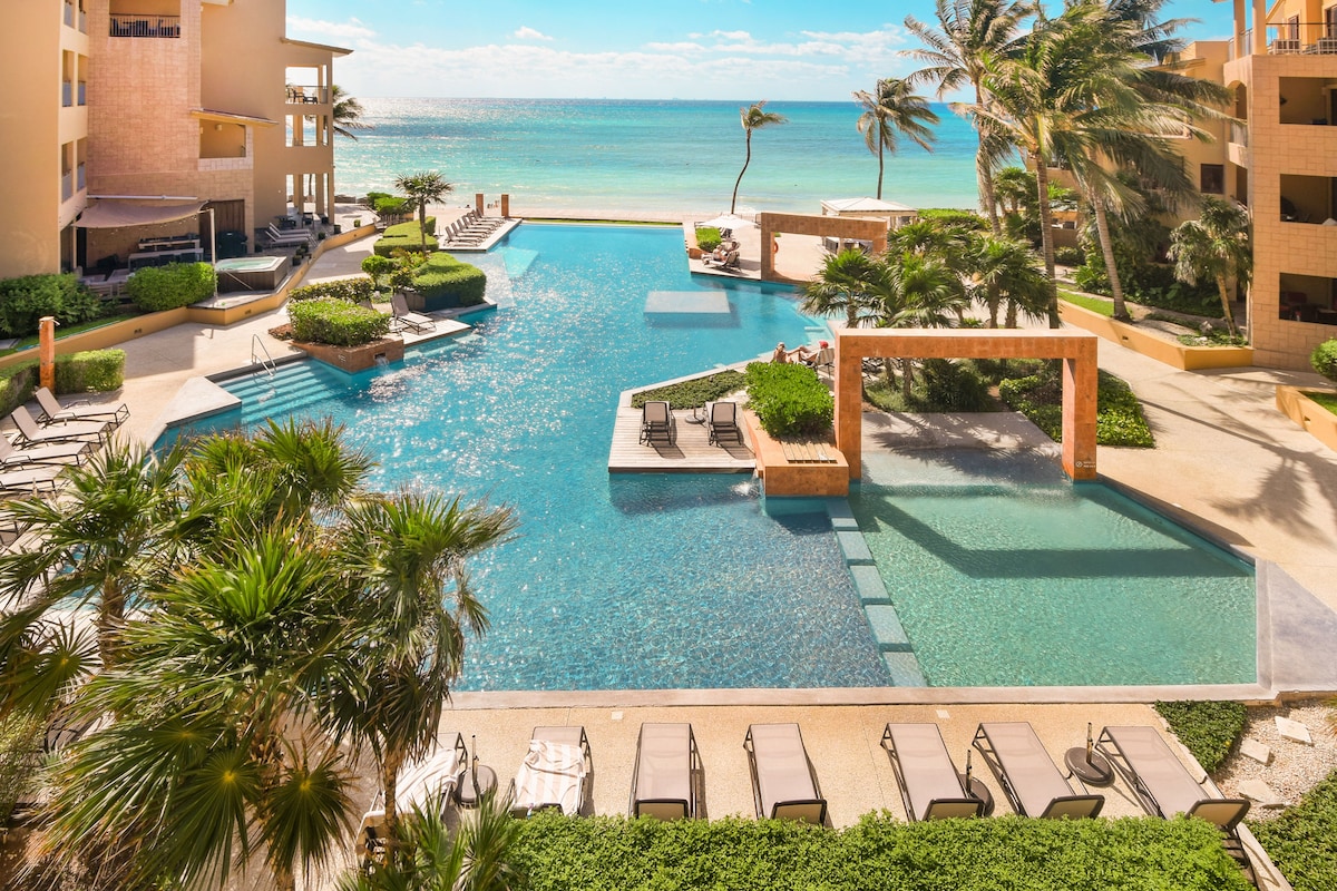 An inviting infinity pool is set against the backdrop of the Caribbean Sea. Resort-style loungers surround the water, and palm trees sway gently in the breeze. The bright blue sky complements the serene atmosphere of the outdoor space.