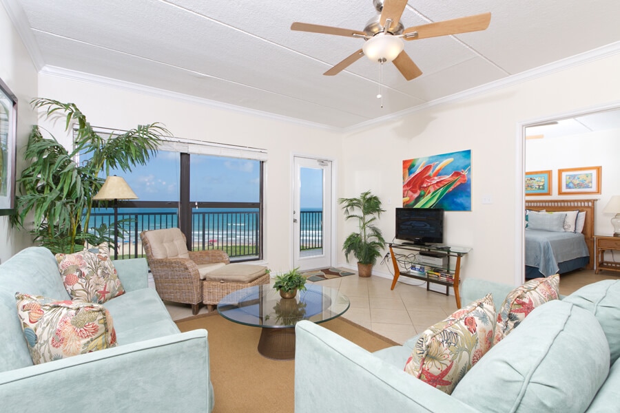 A bright living area is furnished with light blue sofas, a round glass coffee table, and indoor plants. Large windows offer a view of the ocean beyond, while decorative artwork adds a colorful touch. Natural light enhances the airy atmosphere of the space.