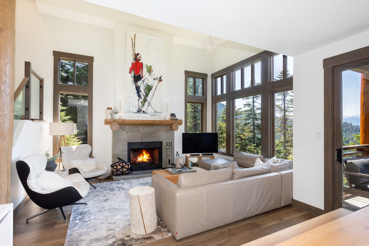 A spacious living area is highlighted by a stone-faced fireplace, with a large piece of art featuring a skier mounted above it. Two comfortable couches and an armchair surround a wooden table, while oversized windows frame views of surrounding trees.