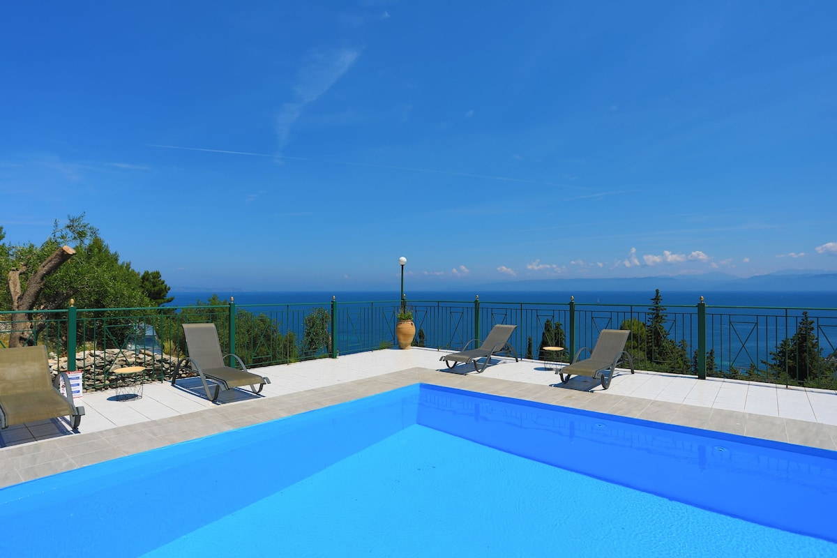 A clear blue pool is positioned against a backdrop of panoramic sea views. Sun loungers are arranged along the poolside, inviting relaxation. Green trees and distant hills are visible beyond the pool, framed by a bright blue sky.
