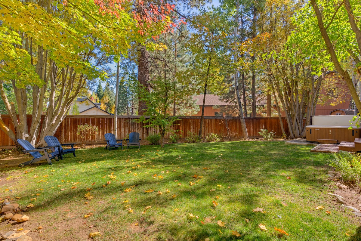 A fenced backyard is depicted, featuring well-maintained grass surrounded by vibrant trees. Several blue adirondack chairs are arranged on the lawn, and a hot tub can be seen at the edge of the space, providing opportunities for relaxation in a serene setting.