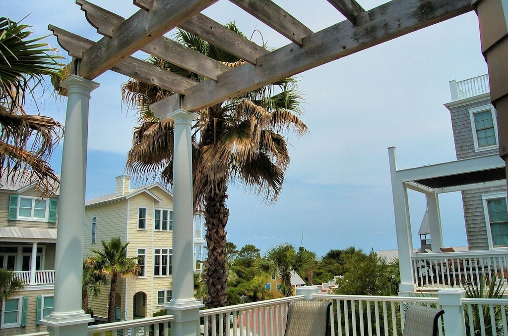 The image captures a view from a deck featuring white railings and a wooden pergola. Lush palm trees and neighboring pastel-colored homes are visible in the background, creating a serene coastal environment. The sky is clear, enhancing the tranquil setting.