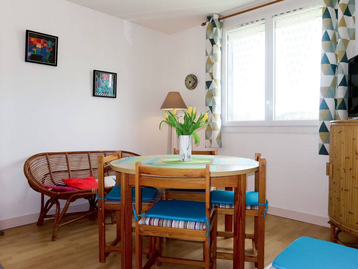 A dining area features a round table surrounded by wooden chairs with bright blue cushions. A rattan bench adds seating, while a vase with yellow tulips serves as a centerpiece. Colorful wall art and patterned curtains complement the natural light streaming through a window.