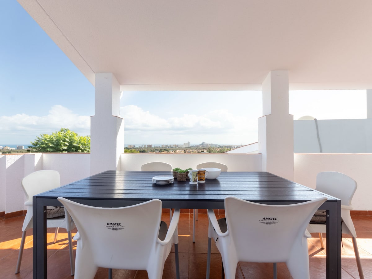 A dining table with six white chairs is situated on a spacious terrace. A scenic view of the sea and city can be observed in the background. The area is illuminated by natural light, creating an inviting atmosphere for outdoor dining.