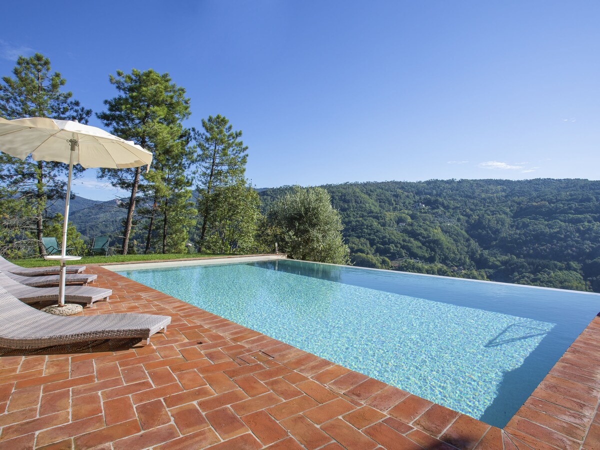 An infinity pool with a tranquil water surface reflects the clear blue sky. Surrounding trees and a scenic view of the hills create a serene backdrop. A sun umbrella and lounge chairs are positioned on the terracotta deck, inviting relaxation.