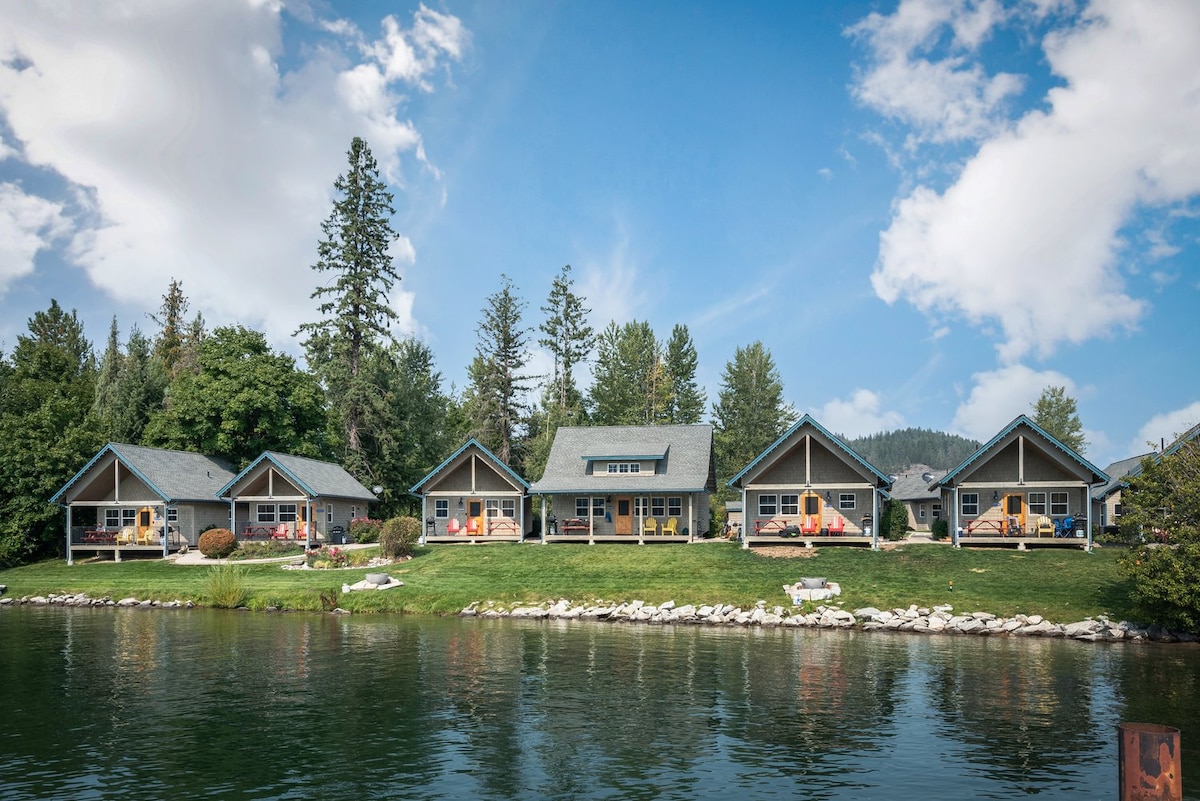 A row of charming bungalows is visible along the water's edge, each adorned with colorful furniture on private porches. Lush greenery and trees provide a natural backdrop, while blue skies with fluffy clouds reflect on the serene lake in front.